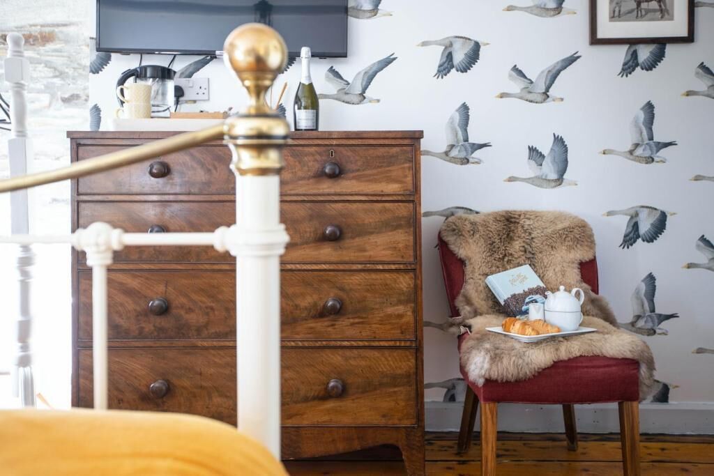 A bedroom with a wooden dresser, a red chair with a fur throw, and a wall with seagull wallpaper.