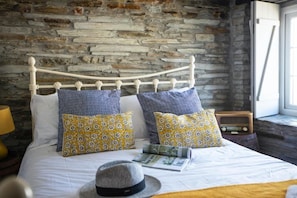 Un lit avec un mur en pierre en arrière-plan, une tête de lit en bois et des coussins à motifs.