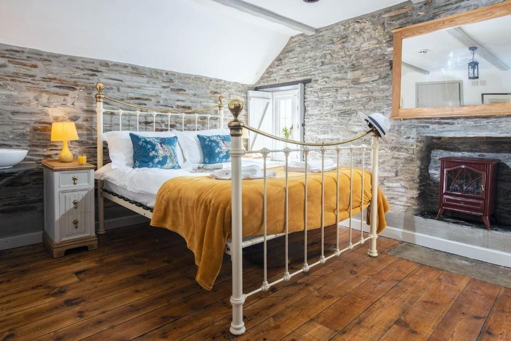 Un dormitorio con pared de piedra, cama con estructura de metal blanco, suelo de madera, mesita de noche con lámpara y chimenea.