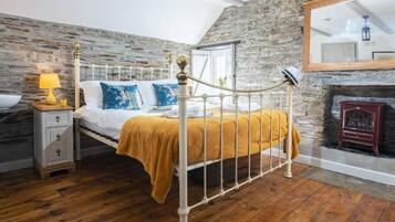 A bedroom with a stone wall, a bed with a white metal frame, a wooden floor, a nightstand with a lamp, and a fireplace.