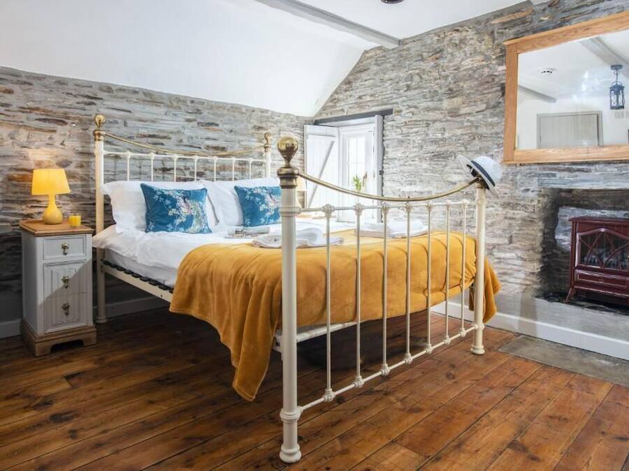 A bedroom with a stone wall, a bed with a white metal frame, a wooden floor, a nightstand with a lamp, and a fireplace.