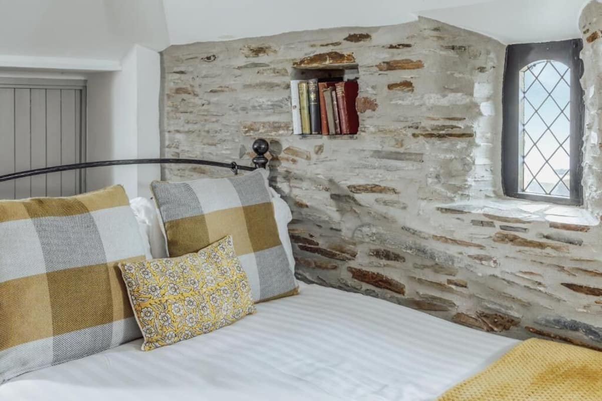 Un dormitorio con pared de piedra, cama con almohadas a cuadros y florales, estantería con libros y ventana con vistas.