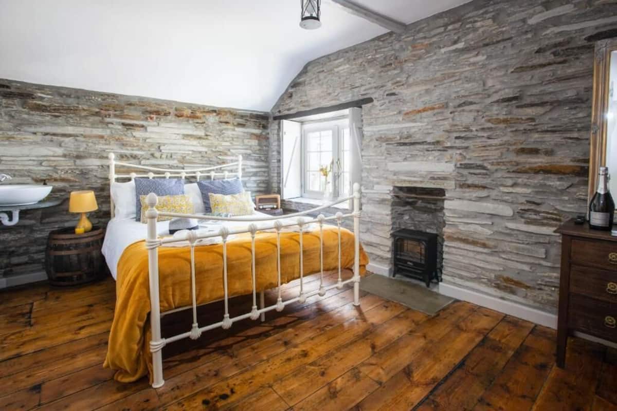 Un dormitorio con pared de piedra, cama con estructura de metal blanco, suelo de madera, ventana y chimenea.