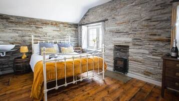 Un dormitorio con pared de piedra, cama con estructura de metal blanco, suelo de madera, ventana y chimenea.