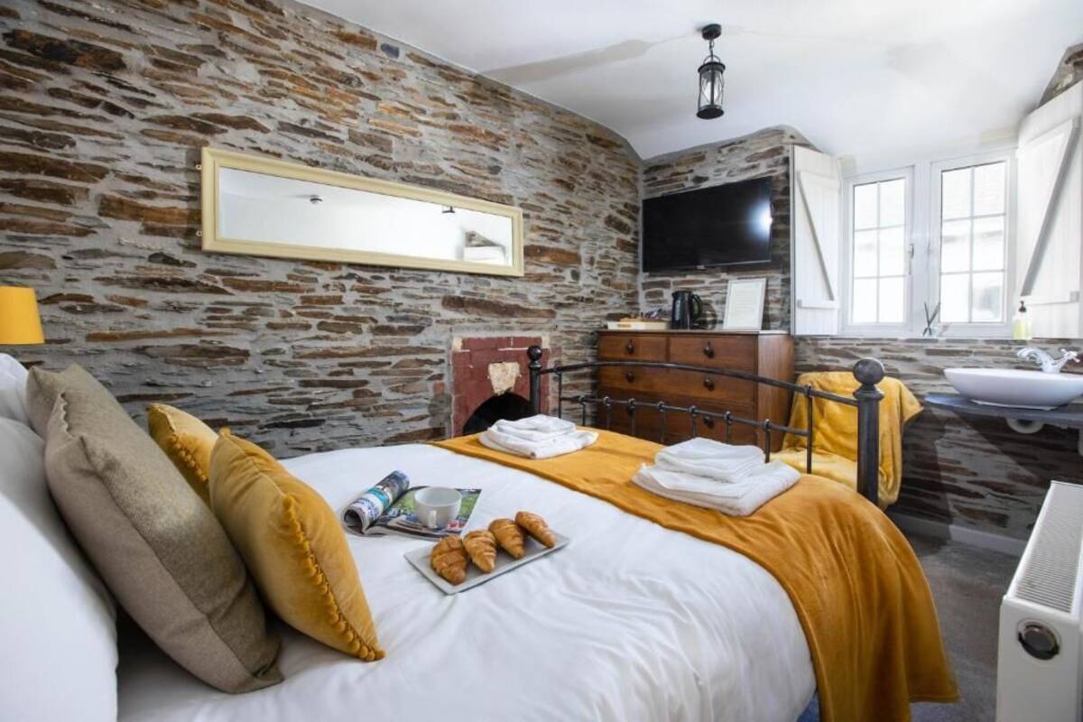 Un dormitorio con pared de piedra, una cama con almohadas, una mesita de noche con lámpara, un televisor y un baño con lavamanos y espejo.