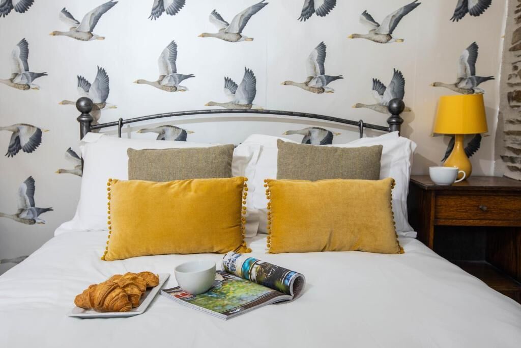 A bed with white bedding and two mustard pillows, a bedside table with a lamp, a cup, and a plate with croissants.