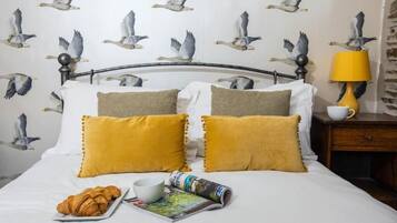 A bed with white bedding and two mustard pillows, a bedside table with a lamp, a cup, and a plate with croissants.