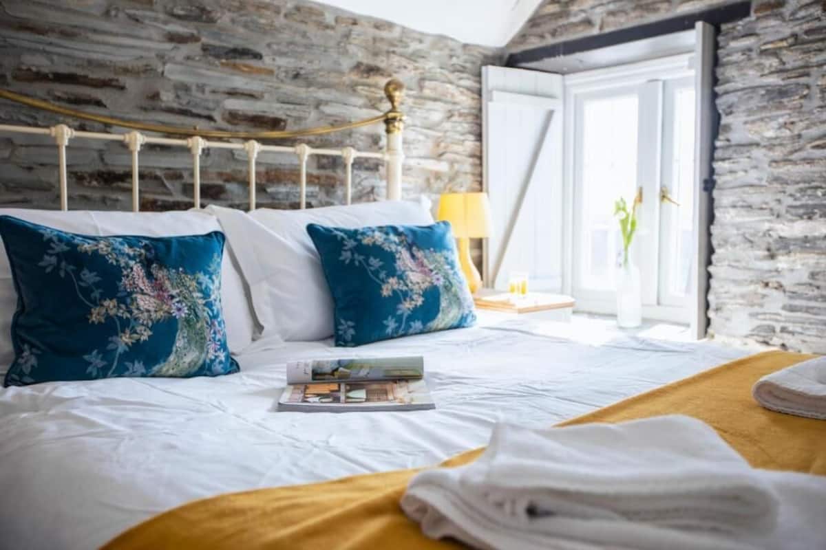 Una cama con ropa de cama blanca, almohadas con estampado floral azul, un cabecero de madera, una mesita de noche con lámpara, y vistas a un muro y una puerta de piedra.