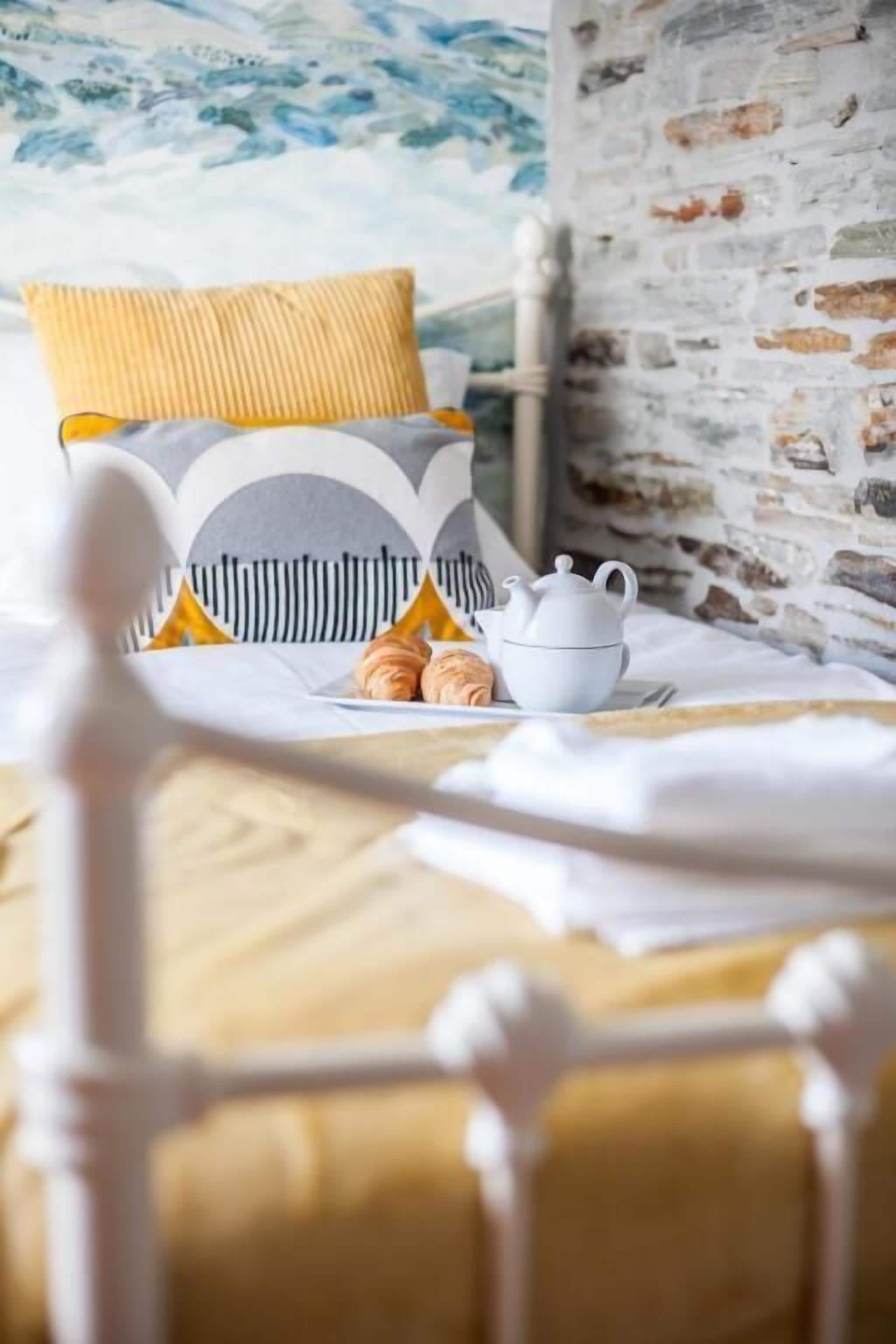 Una cama con cabecera blanca, un cojín amarillo y una tetera blanca con una taza sobre una mesa cercana.
