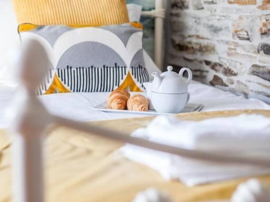 Una cama con cabecera blanca, un cojín amarillo y una tetera blanca con una taza sobre una mesa cercana.