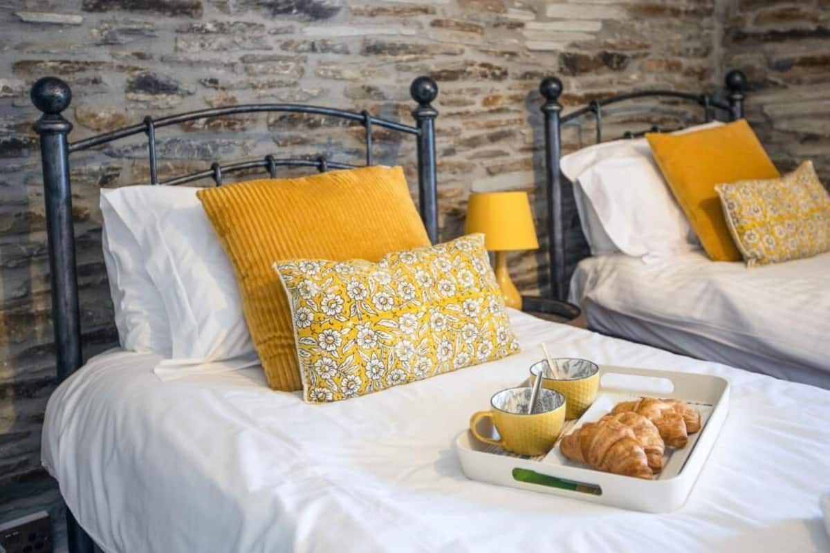 Dos camas con estructura metálica, ropa de cama blanca, almohadas amarillas y estampadas, una bandeja con cruasanes y una taza, y una lámpara en una mesita de noche.