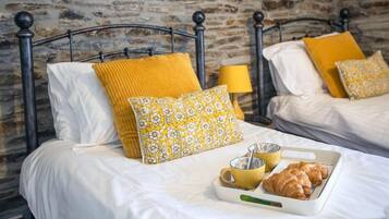 Two beds with metal frames, white bedding, yellow and patterned pillows, a tray with croissants and a cup, and a lamp on a nightstand.