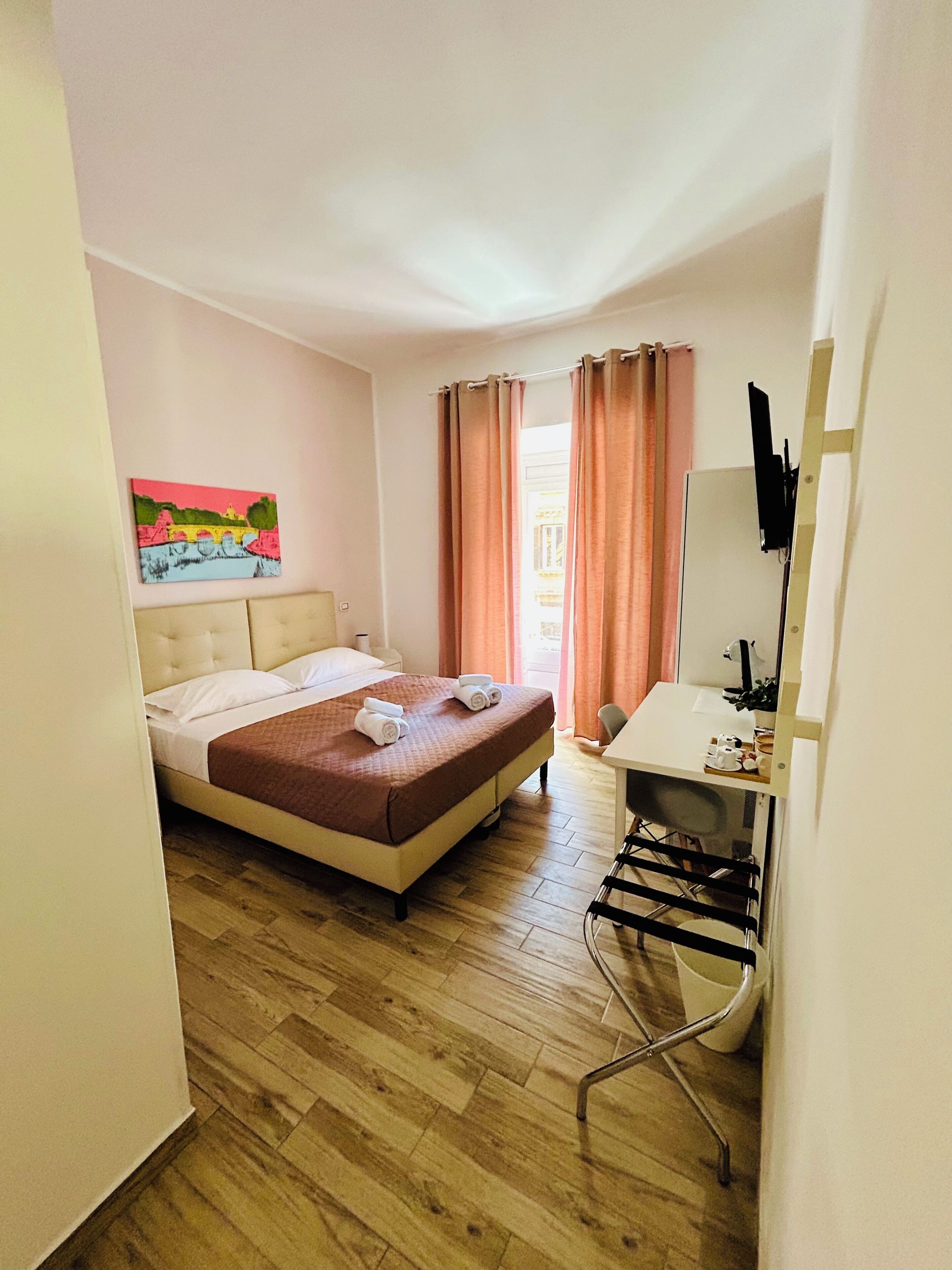 A hotel room with a bed, a desk, a chair, a TV, and a painting on the wall.