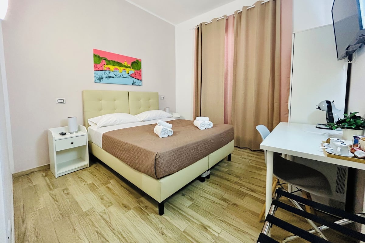 Ein Hotelzimmer mit Bett, Schreibtisch, Sessel, Fernseher, einem Bild an der Wand und einem Fenster mit Vorhängen.