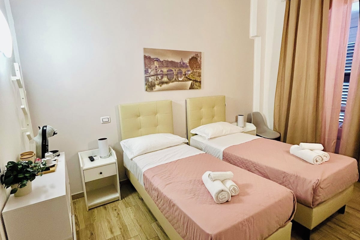 Ein Hotelzimmer mit zwei Betten, einem Schreibtisch, einem Stuhl, einem Nachttisch, einem Bild an der Wand und einem Fenster mit Vorhängen.