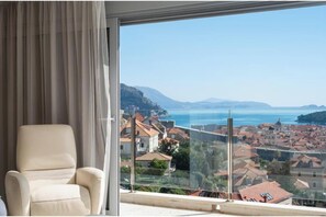 Ein Zimmer mit großem Fenster und Blick auf eine Küstenstadt, ein beiges Sessel und ein Glasgeländer.