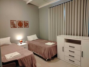 Un dormitorio con dos camas, una mesita de noche con lámpara, un escritorio con cajones y una ventana con persianas.