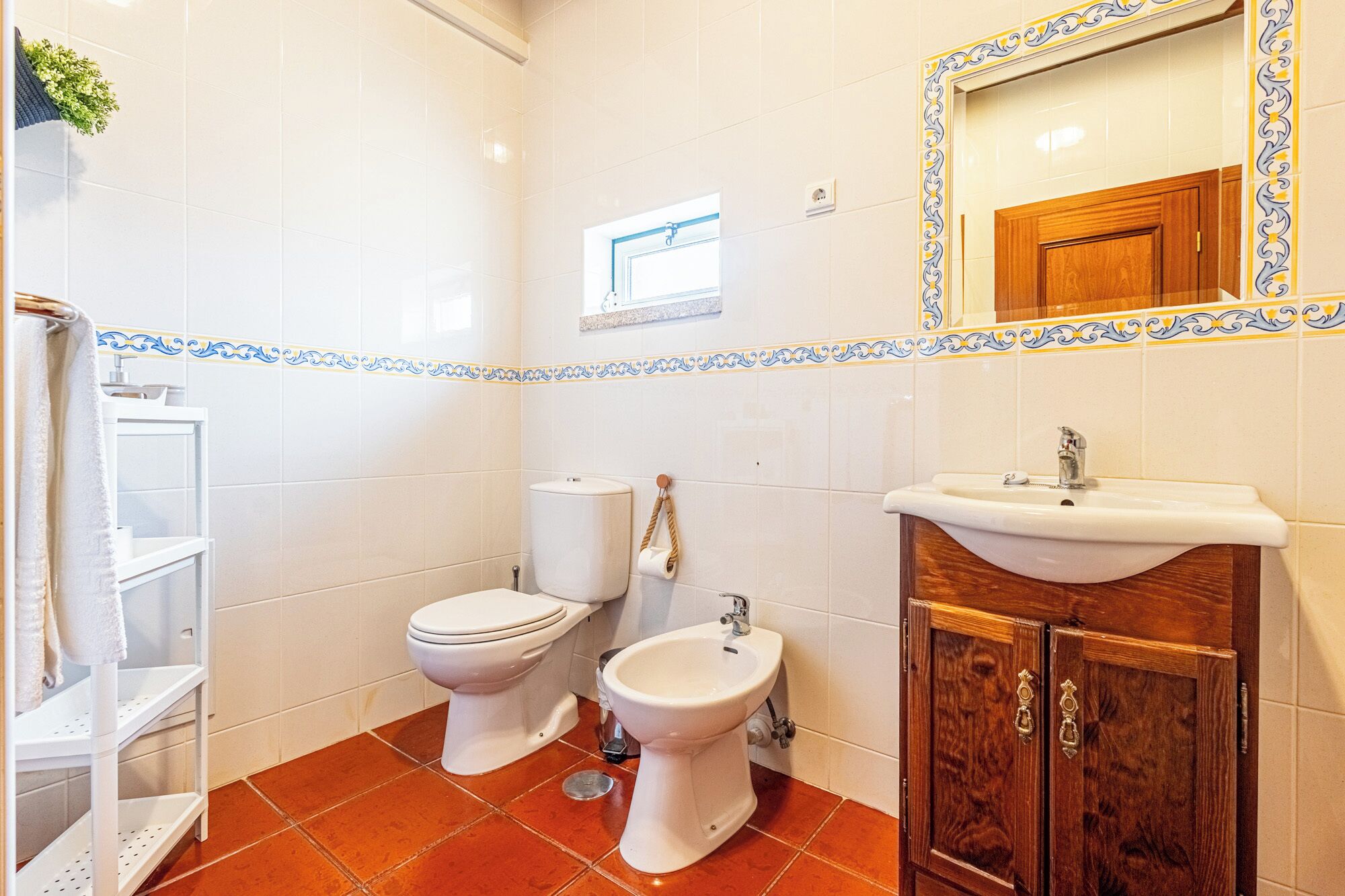 A bathroom with white tiles, a wooden cabinet, a sink, a toilet, and a bidet.