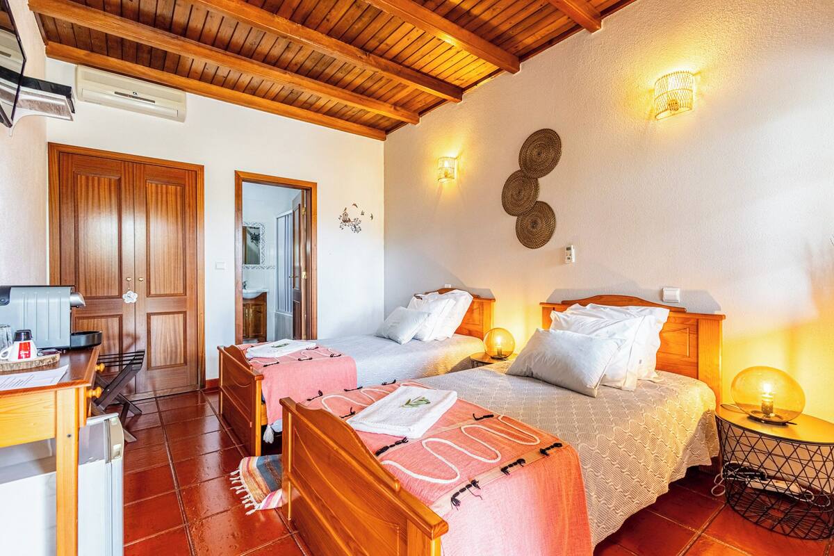 A room with two beds, wooden ceiling beams, a small table with a lamp, and a bathroom visible through an open door.