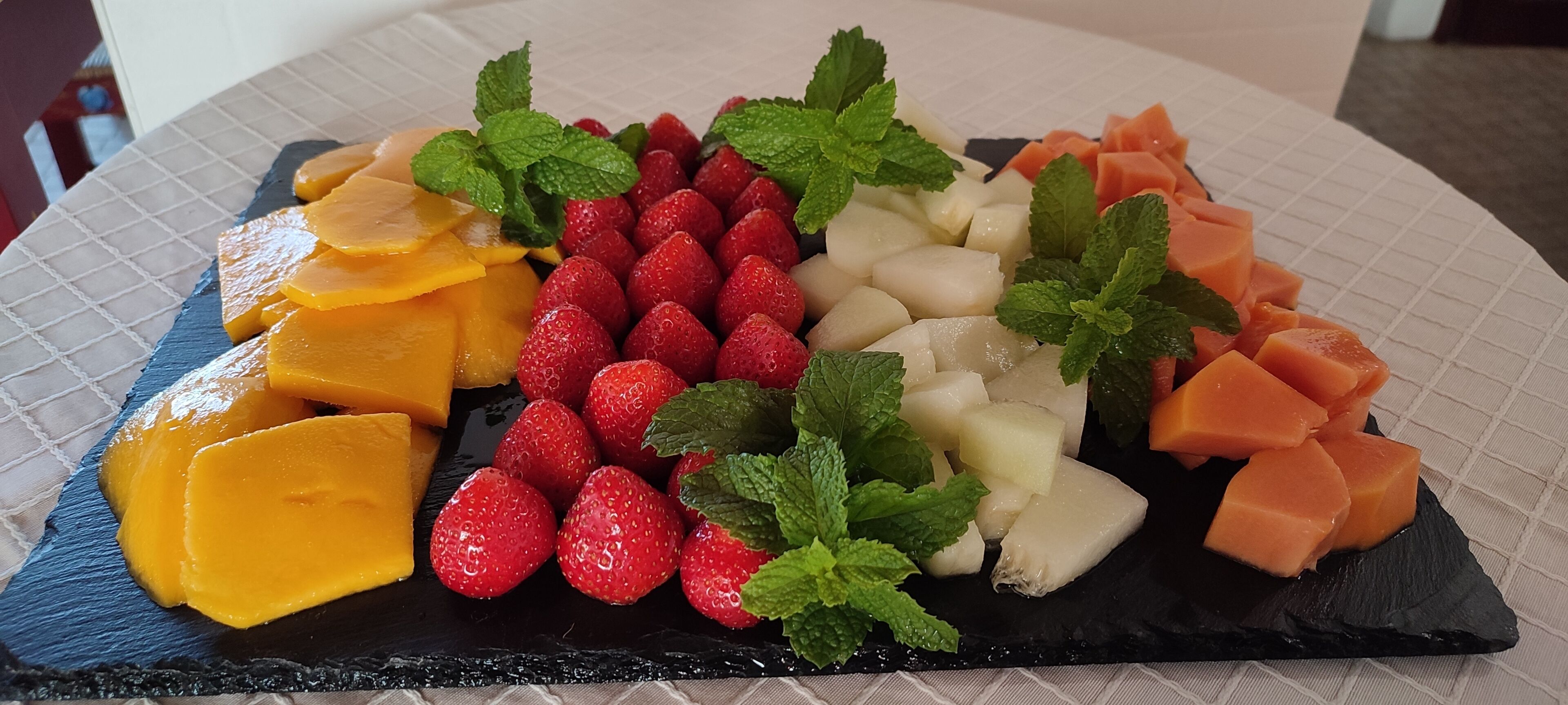 A fruit platter with strawberries, mangoes, and cantaloupe on a black slate plate.