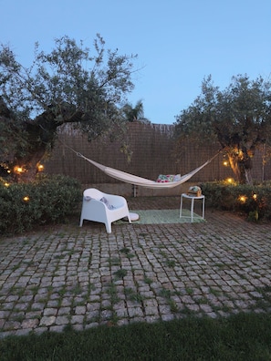 A hammock, a white lounge chair, and a small table in a backyard with a brick floor and a wooden fence. - Monte Além Tejo (Arraiolos)