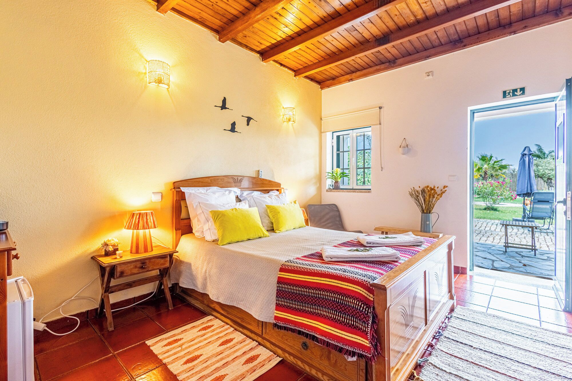 Une chambre avec un plafond en bois, un lit recouvert d’une couverture à motifs, une table de chevet avec une lampe, et une vue sur l’extérieur par une porte.