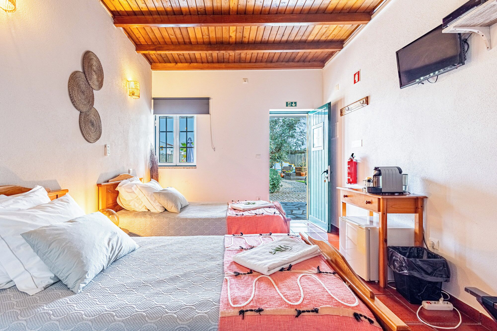 A hotel room with a wooden ceiling, a bed with white bedding, a small table with a kettle, a fire extinguisher, and a TV on the wall.