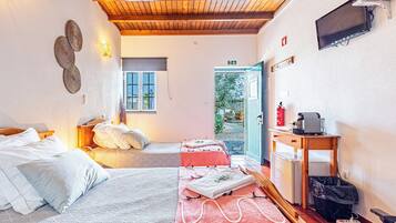 A hotel room with a wooden ceiling, a bed with white bedding, a small table with a kettle, a fire extinguisher, and a TV on the wall.