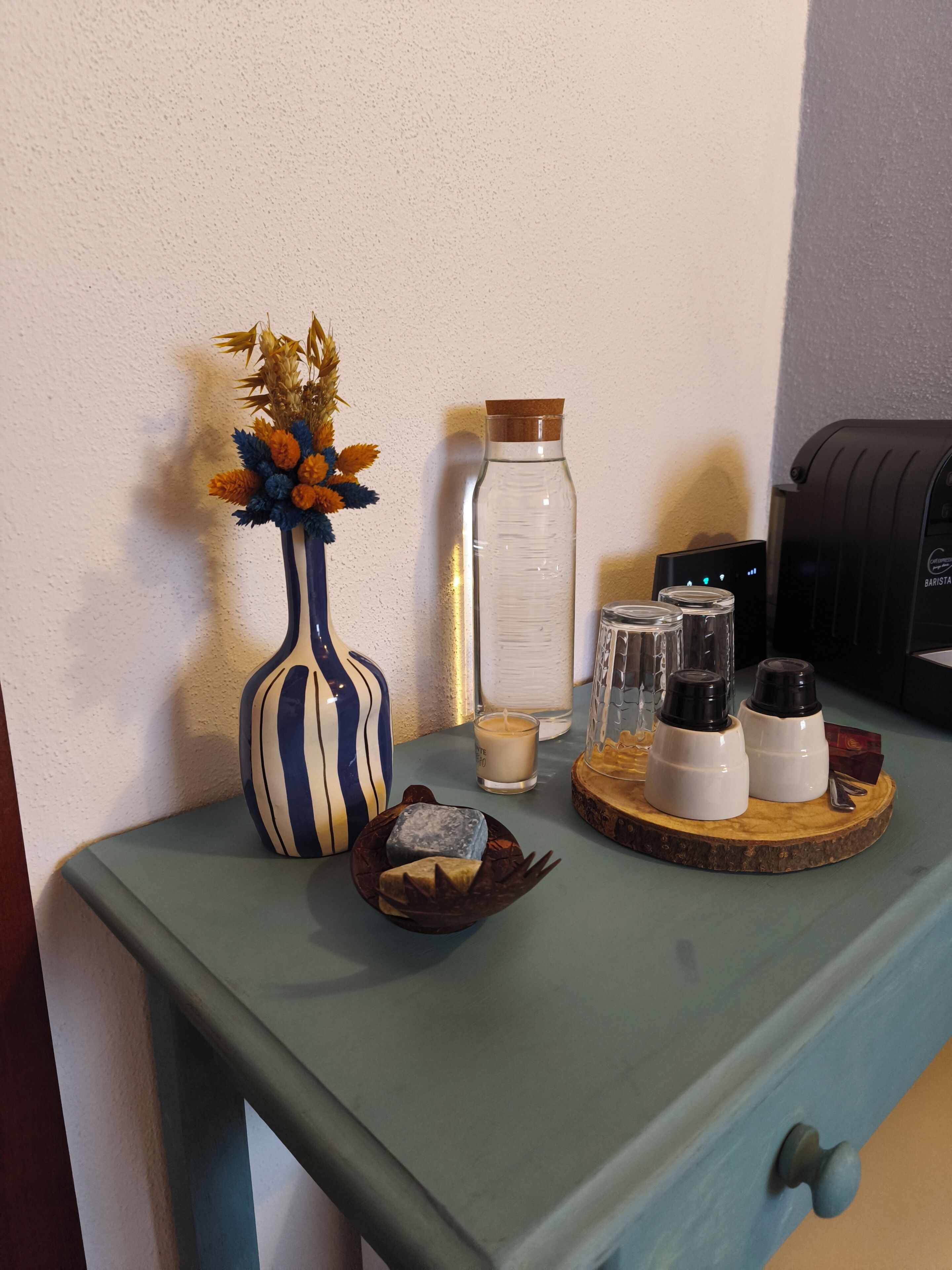 Una estación de barista con una máquina de café, una vela, un florero con flores secas y saleros y pimenteros sobre una bandeja de madera.