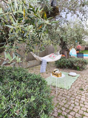 A hammock hanging between trees, a small table with a cushion, and a brick pathway.