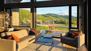 Ein modernes Wohnzimmer mit einer Couch, Sesseln und einem Couchtisch. Große Fenster bieten Blick auf eine grüne Landschaft und Hügel.