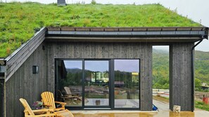 A modern wooden building with large glass doors, outdoor seating, and a view of a green hillside.