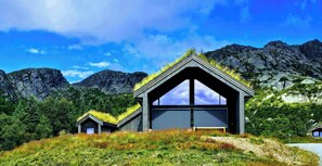Une maison moderne dotée d’une grande baie vitrée au centre et de plus petites fenêtres sur les côtés, entourée d’un paysage verdoyant et de montagnes.