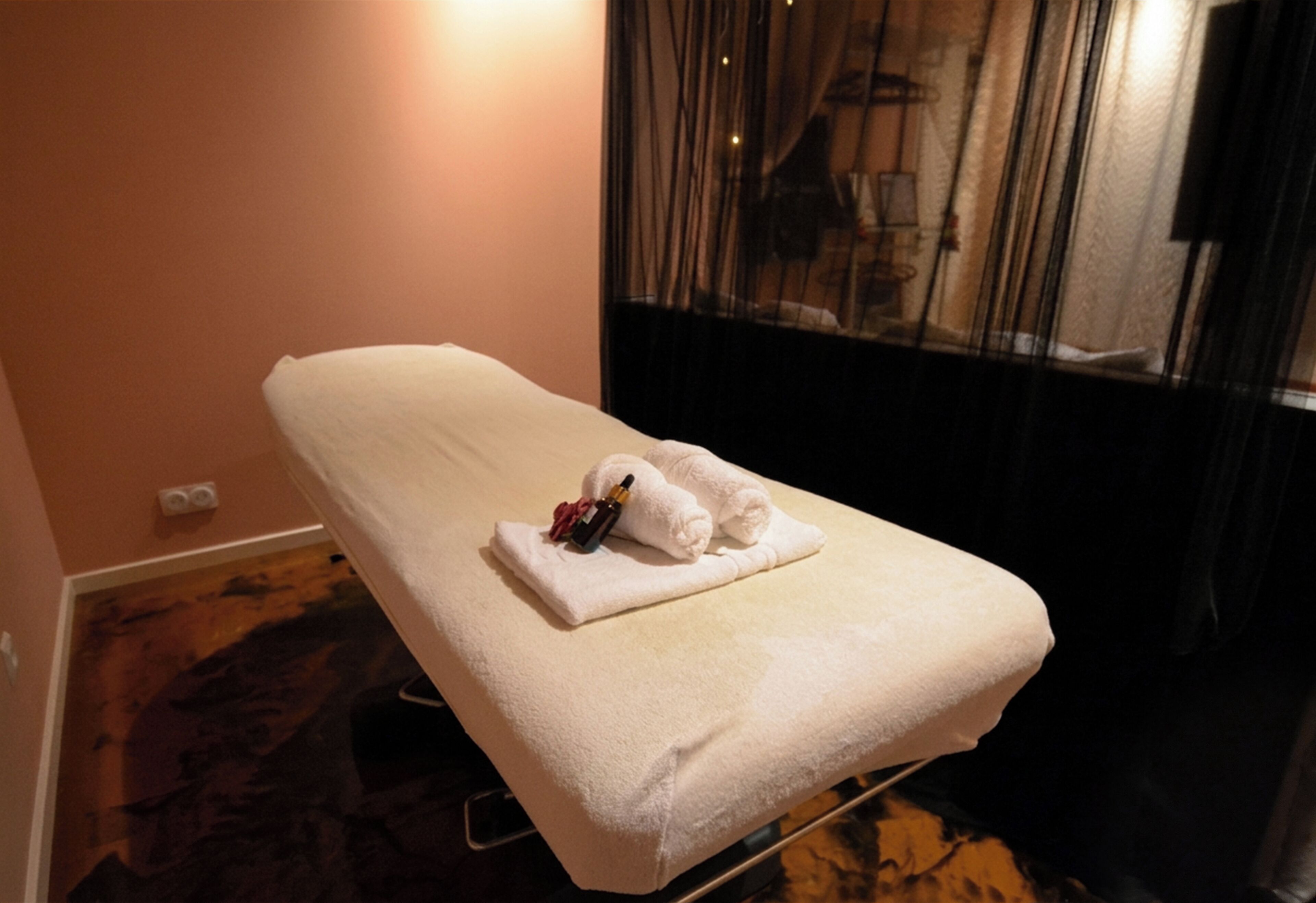 A single bed with a white towel and a bottle, a wall socket, and a window with curtains.