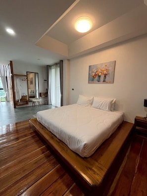 A bedroom with a large bed, wooden flooring, a painting on the wall, and a bathroom visible through an open door. - Mei Nai Home (Pua)