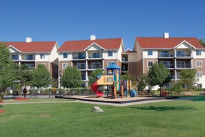 A playground with a slide and climbing structures, surrounded by a well-maintained lawn and a row of multi-story residential buildings with red roofs. - CW BLOCK Mountain Vista 2br (Branson)