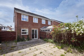 A two-story brick house with a front door, a garage door, and a small front yard with a wooden fence and flowering plants. - Host Stay - Urban Nest LIV (Liverpool)