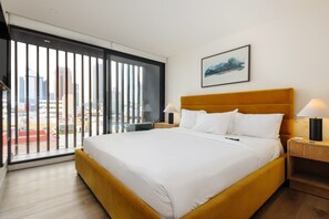 Ein Hotelzimmer mit einem großen Bett, zwei Nachttischen mit Lampen, Blick auf die Stadt durch ein großes Fenster und einem gerahmten Bild an der Wand.