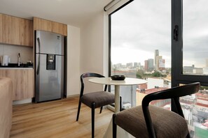 A modern kitchen with a stainless steel refrigerator, wooden cabinets, a round dining table with two chairs, and a large window offering a city view. - w Urban 1BR w/ AC in the Heart of Cuauhtemoc (Mexico City)