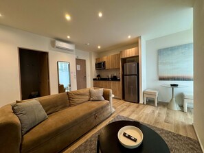 Ein modernes Wohnzimmer mit einer Couch, einem kleinen Tisch mit einer Schüssel, einer Küchenzeile mit Holzschränken, einem Kühlschrank und einem Fenster mit Blick.