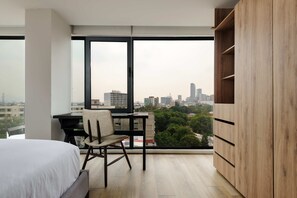 Ein Schlafzimmer mit Bett, Schreibtisch und Stuhl, einem großen Fenster und Blick auf eine Stadtlandschaft.