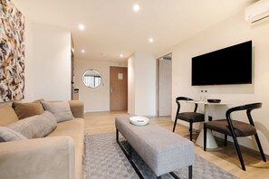 Ein modernes Wohnzimmer mit einer Couch, einer Bank, einem Esstisch mit Stühlen, einem Flachbildfernseher und einem runden Spiegel.