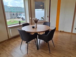 A room with a round wooden table, four chairs, a radiator, a window with a view of a building and parked cars, and a plant on the windowsill. - Ferinhaus near the coast (Großenwiehe)