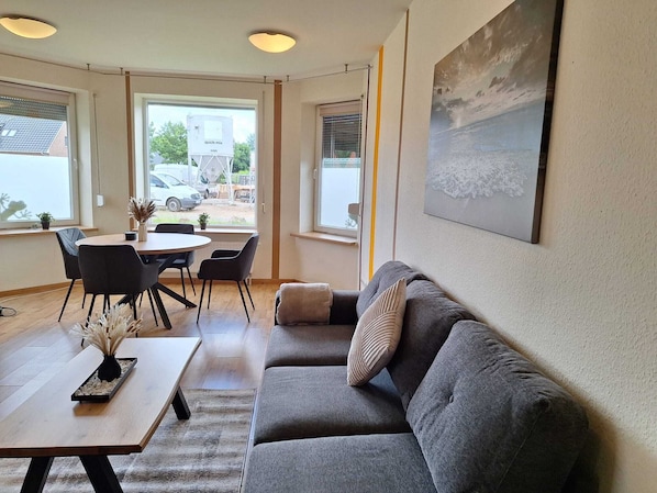 Un salon moderne avec un canapé gris, une table ronde et des chaises, de grandes fenêtres donnant sur l’extérieur et un tableau accroché au mur.