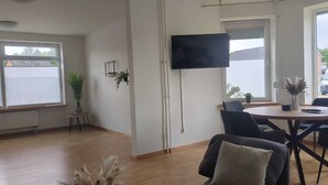 Ett modernt vardagsrum med en platt-TV som är monterad på väggen, ett runt matbord med stolar, en krukväxt och ett fönster med persienner.
