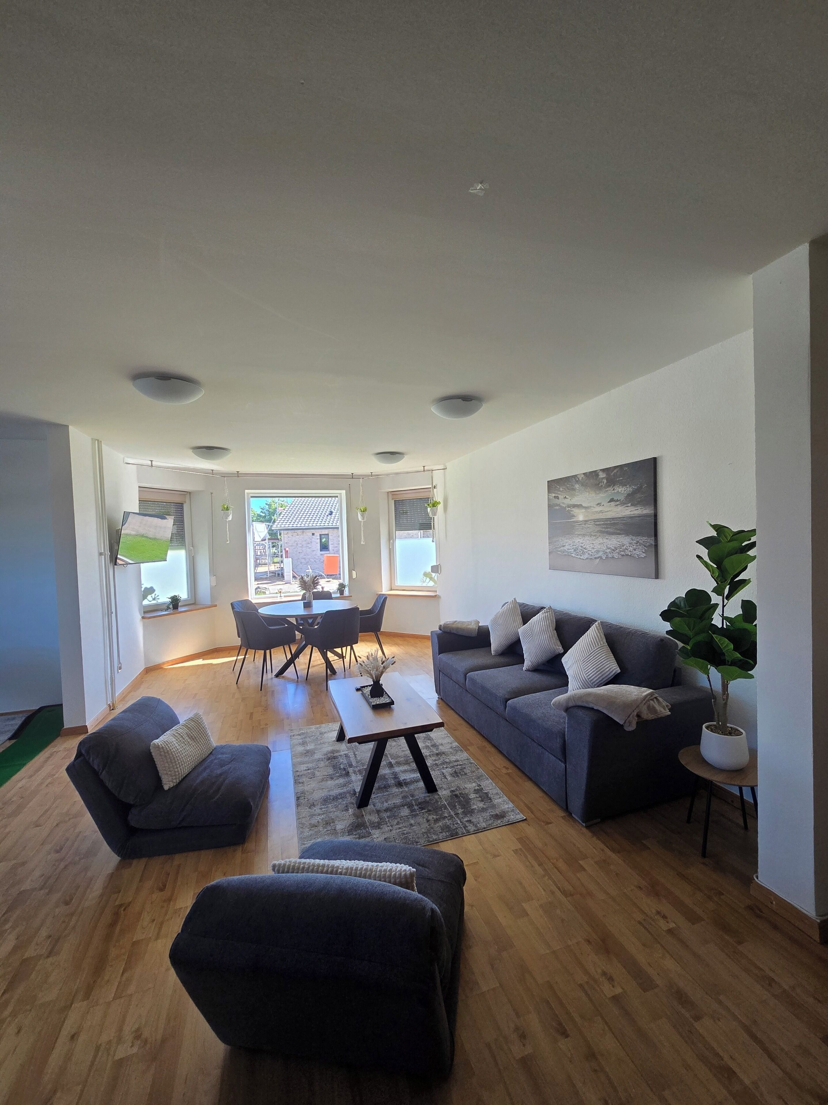 Una sala moderna con sofá, reposapiés y mesa de centro, un comedor con sillas y una mesa, y un ventanal con vistas.