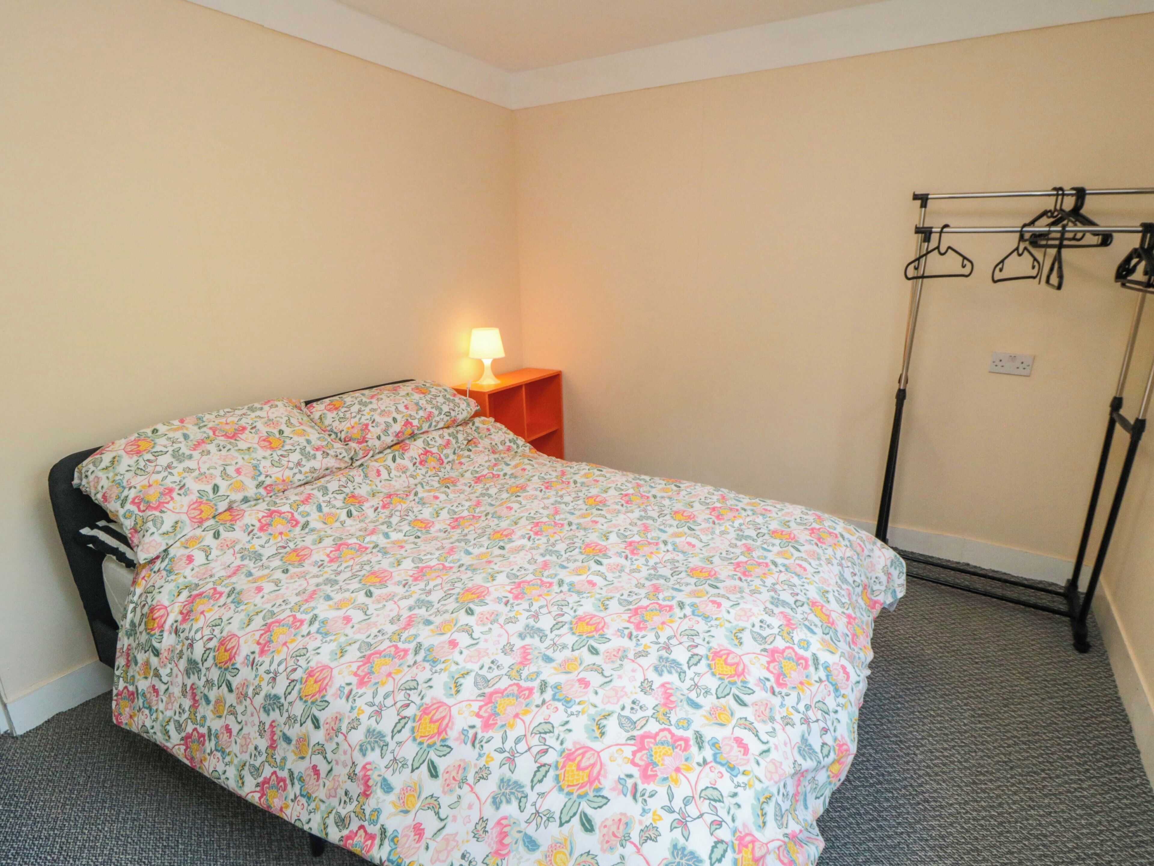 A bedroom with a floral bedspread, a bedside table with a lamp, and a clothing rack.