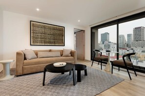 Ein modernes Wohnzimmer mit einer Couch, zwei Tischen, Stühlen, einem großen Fenster mit Blick auf die Stadt und einem gerahmten Kunstwerk an der Wand.