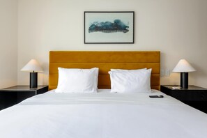 Ein Hotelzimmer mit einem Bett, zwei Nachttischen, einem kleinen Tisch, einem gerahmten Kunstwerk und einer Fernbedienung auf dem Bett.