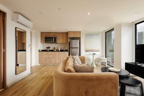 Ein modernes Wohnzimmer mit Sofa, Couchtisch und Fernseher. Es gibt eine Küchenzeile mit Holzschränken und einem Kühlschrank.